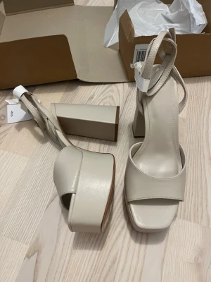 Beige platåsandaler med blockklack, 38, Mango - Ny Snygga beige sandaler från Mango med hög platå och bred blockklack, storlek 38.  Skorna har fyrkantig tå, bred rem över foten och justerbar ankelrem. Tillverkade i slätt konstläder som ger en stilren look. Perfekta för dig som vill ha extra höjd och trendig stil.