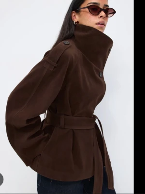 Mörkbrun trenchcoat med bälte - Snygg mörkbrun trenchcoat från Gina med hög krage, breda ärmar och knytskärp i midjan. Jackan har dekorativa axelklaffar och stora knappar framtill. Tillverkad i ett mjukt tyg som ger en lyxig känsla och passar perfekt till jeans eller byxor. Helt ny❤️