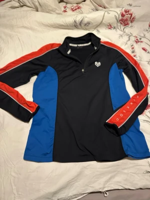 Svart och blå sporttröja från pink soda - Sportig långärmad tröja i svart med blå sidopaneler och röda detaljer längs ärmarna. Tröjan har half zip framtill och logga på bröstet. Perfekt för träning eller utomhusaktiviteter.