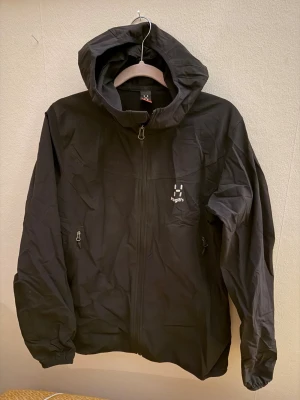 Svart vindjacka från Haglöfs - Snygg svart vindjacka från Haglöfs med dragkedja framtill, två sidofickor med dragkedja och justerbar huva. Jackan har en minimalistisk design med Haglöfs-logga på bröstet. Perfekt för blåsiga dagar och outdoor-äventyr.Står women L men passar mer S-M (min son har haft den)