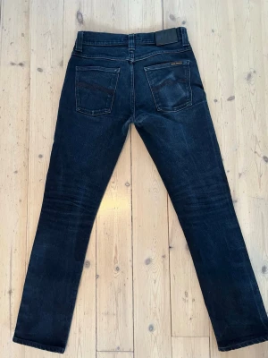 Mörkgrå dry denim jeans från nudie - Snygga mörkgrå/svarta jeans från Nudie Jeans i modellen Grim Tim med rak passform och klassisk femficksdesign. Färgen heter Black Ring och har en snygg, något tvättad finish som ger en stilren och modern look. Jeansen har subtila slitningar och naturliga märken som ger karaktär. Inga hål eller defekter, endast mindre märken från plånbok på bakfickan samt några små bruksspår. Storlek 30W/32L. Osäker på om det är dry denim, men märkena tyder på att de kan vara det. Skriv gärna vid frågor. Mvh Olof