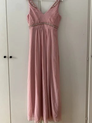 Balklänning med pärldetaljer - Ljusrosa långklänning med v-ringning. Pärlderaljer och guldiga broderier vid midjan och axelbanden. Ytter och innertyg i 100% polyester.