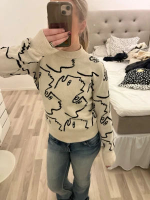 Beige stickad tröja med ansiktsmönster - Trendig beige stickad tröja med svart abstrakt ansiktsmönster över hela plagget. Tröjan har ribbad hals samt en avslappnad passform. Perfekt för dig som gillar unik design och vill sticka ut. Använd 2 gånger tidigare.