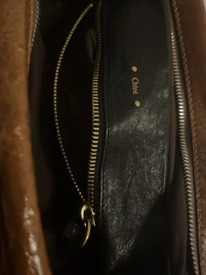 Brun handväska från Chloé i läder - Snygg brun handväska från Chloé i glansigt läder med guldfärgade dragkedjor och detaljer i guld. Väskan har dubbla handtag och rymligt huvudfack med innerficka som stängs med dragkedja. Perfekt för dig som gillar klassisk men väldigt prydlig  design. Mycket bra skick och nästan aldrig använt förutom 2 gånger 