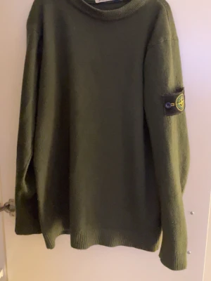 Mörkgrön stickad tröja Stone Island - Säljer en mörkgrön stickad tröja från Stone Island med klassisk patch på vänster ärm. Tröjan har rund halsringning, ribbade muddar och är tillverkad i mjukt material som passar perfekt för kyligare dagar.