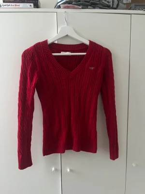 Röd kabelstickad tröja Hollister - Snygg röd kabelstickad tröja från Hollister med V-ringning och lång ärm. Tröjan har en liten broderad logga på bröstet och ribbade muddar vid ärmslut och nederkant. Perfekt för dig som gillar klassisk stickad stil.
