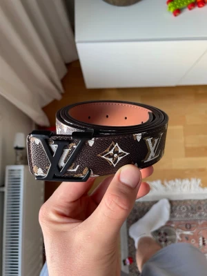 Lv bälte - Säljer ett svart skärp från Louis Vuitton med det klassiska LV-monogrammet i silver och guld på hela bandet. Bältet har en bred design och är gjort i skinn med exklusiv känsla. Perfekt för dig som vill ha en statement-accessoar.