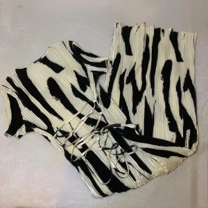 Zebramönstrad klänning med snörning - Cool zebramönstrad klänning i svart och vitt med korta ärmar och trendig snörning i midjan. Plisserat tyg ger en snygg struktur och en unik look. Perfekt för dig som vill sticka ut med ett djurmönster och en modern passform.