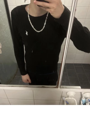 Svart stickad tröja från Polo Ralph Lauren - Klassisk svart stickad tröja från Polo Ralph Lauren med rund halsringning och vit broderad logga på bröstet. Tröjan har ett stilrent kabelstickat mönster och långa ärmar. Perfekt att matcha med jeans eller chinos.