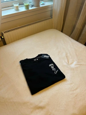 Svart Stüssy x Nike t-shirt - Svart t-shirt från Stüssy i samarbete med Nike. Framsidan har stora vita tryck med Stüssy-logga, ett peace-märke, ett hjärta och Nike swoosh. Klassisk passform och rund hals. Perfekt för dig som gillar streetwear och vill sticka ut.