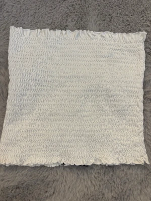 Vit tubtopp från Bik Bok - Säljer en vit bandeau tubtopp från Bik Bok med smockad struktur och vågiga kanter. Toppen är ärmlös och har en stretchig passform som sitter snyggt på kroppen. Perfekt att styla till jeans eller kjol för en fräsch look. 