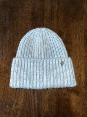 Ljusblå ribbstickad mössa - Mysig ljusblå ribbstickad mössa med uppvikt kant och liten metalllogga på sidan. Tillverkad i mjukt garn som ger en fluffig känsla och håller dig varm under kalla dagar. Perfekt accessoar för att lyfta vinterlooken.