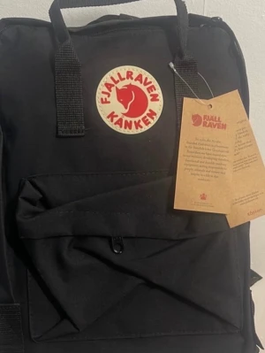 Svart Fjällräven Kånken ryggsäck - Klassisk svart Fjällräven Kånken ryggsäck med det ikoniska runda röda och vita logotypmärket framtill. Ryggsäcken har ett stort huvudfack, en framficka med dragkedja och bärhandtag upptill. Tillverkad i slitstarkt syntetmaterial. 16L storlek