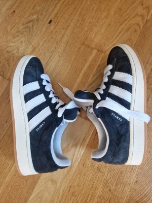 Adidas Campus svarta sneakers - Snygga Adidas Campus sneakers i svart mocka med klassiska vita ränder på sidorna och vit sula. Skorna har vita skosnören och en retroinspirerad design som passar perfekt till streetwear. Yttersulan är i gummi med bra grepp.