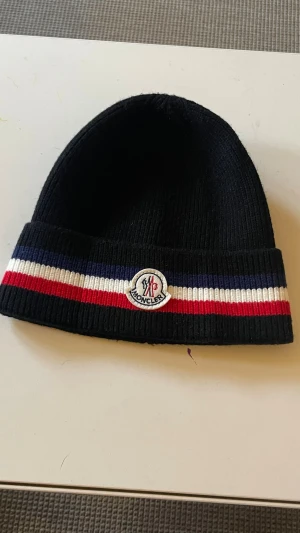 Svart Moncler mössa med ränder - Säljer en svart ribbstickad mössa från Moncler med bred uppvikt kant. Mössan har klassiska Moncler-loggan framtill och snygga ränder i blått, vitt och rött runt om. Perfekt accessoar för dig som vill ha en stilren och trendig look.