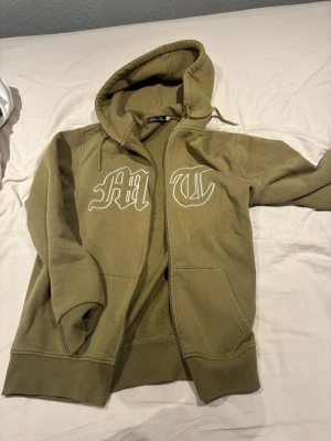 Hoodie från Tuttolente  - Säljer en olivgrön hoodie från Tuttolente med dragkedja och stora vita bokstäver på bröstet. Hoodien har huva med dragsko, ribbade muddar och mjukt fleecefoder på insidan. Perfekt för en chill och avslappnad stil. Passar även storlek M.