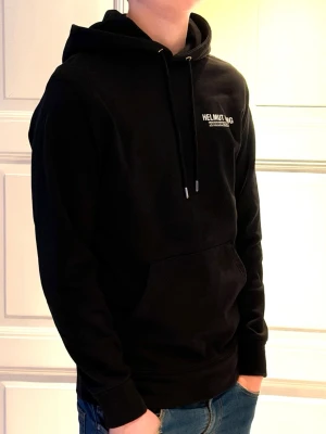 Svart hoodie från Helmut Lang - (Moddelen är 185-186 och väger 75 kg)Snygg svart hoodie från Helmut Lang med vit logga på bröstet. Klassisk känguruficka framtill, justerbar dragsko i huvan och ribbade muddar vid ärmar och midja. Tillverkad i mjukt material som känns skönt mot huden. Perfekt för en avslappnad och trendig look.