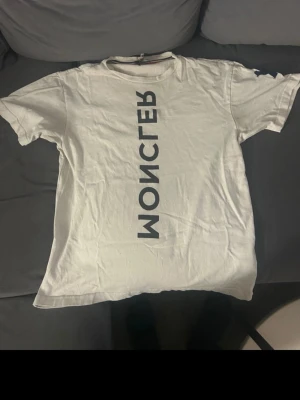 Vit Moncler t-shirt med tryck - Säljer en vit t-shirt från Moncler Grenoble med ett stort vertikalt tryck på framsidan. Klassisk passform och rund halsringning. Tillverkad i mjuk bomull som känns skön mot huden. Perfekt för dig som gillar stilrena och exklusiva plagg.