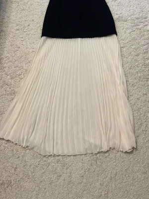 Svart och vit långklänning med plisserad kjol - Snygg långklänning med svart överdel samt vit plisserad kjol. Klänningen har dragkedja i bak och en elegant tvåfärgad design som ger en stilren look. Perfekt för dig som vill sticka ut med en klassisk men modern stil.