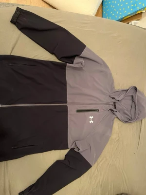 Grå och svart vindjacka Under Armour - Snygg tvåfärgad vindjacka från Under Armour i grått och svart. Jackan har huva, hel dragkedja framtill, två sidofickor med dragkedja och en bröstficka med dragkedja. Tillverkad i lätt och slitstarkt material, perfekt för blåsiga dagar.