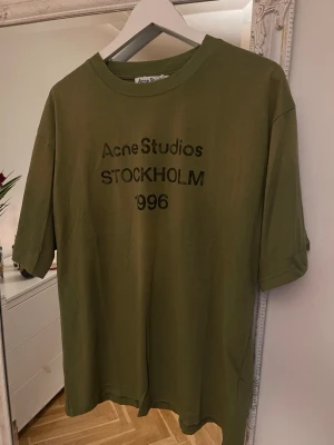 Olivgrön Acne Studios t-shirt - Snygg olivgrön t-shirt från Acne Studios med tryck 'Acne Studios STOCKHOLM 1996' på bröstet. Klassisk rund halsringning och avslappnad passform. Perfekt till jeans eller shorts för en clean och trendig look.