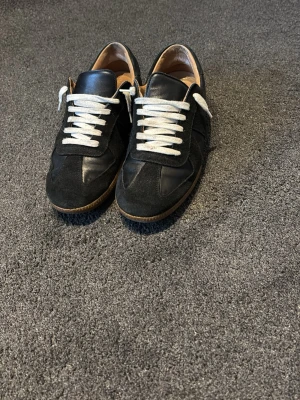 Gats - Snygga svarta sneakers med vita snören och bruna gummisulor. Skorna har en mix av mocka och slätt läder på ovandelen, vilket ger en stilren och klassisk look. Låg modell som passar perfekt till jeans eller chinos.