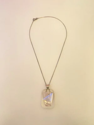 Halsband med transparent hänge - Unikt halsband med ett rektangulärt transparent hänge som har en innesluten metallisk detalj och små pärlliknande dekorationer. Kedjan är tunn och silverfärgad, vilket ger ett stilrent och modernt intryck. Perfekt för dig som gillar smycken med en twist.