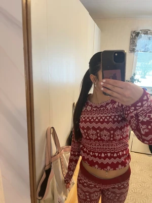 Röd julpyjamas med snöflingor - Mysig röd pyjamas med vitt julmönster och snöflingor. Setet består av en långärmad tröja och matchande byxor med resår i midjan. Perfekt för vinterkvällar och julmys. Materialet ser ut att vara mjukt och stretchigt för extra komfort.