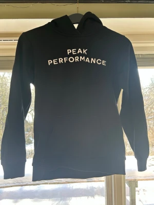 Svart hoodie Peak Performance - Snygg svart hoodie från Peak Performance med vit text på bröstet. Klassisk modell med huva och känguruficka framtill. Perfekt för en avslappnad och sportig stil. Mjuk och skön bomullsblandning som passar året runt.