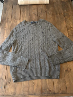 Grå kabelstickad tröja Polo Ralph Lauren - Kabelstickad grå tröja från Polo Ralph Lauren med rund halsringning och diskret broderad logga på bröstet. Tröjan har ribbade muddar vid ärmslut och nederkant, och är tillverkad i mjukt material som passar perfekt till höst och vinter.