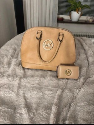 Beige handväska från Michael Kors - Snygg beige handväska från Michael Kors i slätt konstläder med guldfärgad MK-logga framtill. Väskan har två handtag och medföljer en matchande plånbok med samma logga. Perfekt för dig som gillar stilrena accessoarer och vill ha plats för det viktigaste.