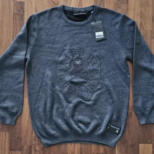 Mörkgrå tröja från Philipp Plein - size XXLarge  - Säljer en mörkgrå tröja från Philipp Plein med broderad döskalle och orm-motiv på bröstet. Tröjan har rund halsringning och långa ärmar. Perfekt för dig som gillar streetwear och exklusiva detaljer.