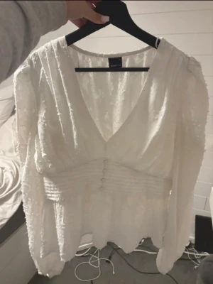 Vit romantisk blus med v-ringning - Säljer en vit blus från Gina Tricot med v-ringning, dekorativa knappar framtill och långärmad design. Blusen har ett lätt strukturerat tyg med diskret mönster och markerad midja för en feminin siluett.