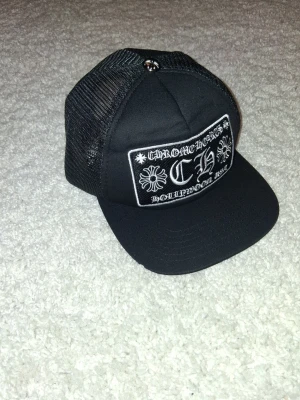 Chrome Hearts truckerkeps svart - Säljer en svart truckerkeps från Chrome Hearts med vit brodyr framtill. Kepsen har mesh på sidorna och bak, samt en böjd skärm. På framsidan står det 'Chrome Hearts Hollywood USA' och har dekorativa korsdetaljer.