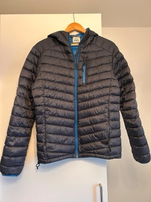 Mörkblå pufferjacka från Mc Kinley - Snygg mörkblå pufferjacka från Mc Kinley med luva och blå dragkedjor. Jackan har bröstficka med dragkedja och två sidofickor. Perfekt för kalla vinter-, höst- och vårdagar. Tillverkad i lätt, värmande material. Storlek XS Herr. 