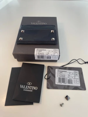 Svart plånbok Valentino Garavani - Snygg svart plånbok från Valentino Garavani med coola silverfärgade nitar på framsidan. Tillverkad i slitstarkt tyg med läderdetaljer och diskret logga. Kommer med originalask, tags och extra nitar. Perfekt för dig som gillar exklusiva accessoarer.