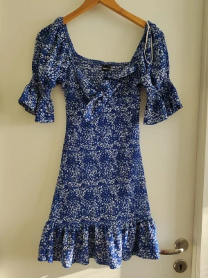 Blommig blå kortklänning Gina Tricot - Supersöt blå kortklänning från Gina Tricot med vitt blommönster, puffärmar och volangkant nertill. Klänningen har en söt knytning framtill och smockad rygg för extra bekväm passform. Perfekt för sommarens alla tillfällen!