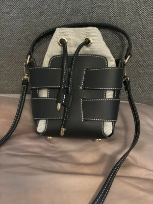 Svart och beige bucket bag med snörning - Trendig bucket bag i svart läder och beige canvas med vita kontrastsömmar och guldfärgade detaljer. Väskan har justerbar axelrem och snörning upptill. Snygg design med rutmönstrade läderband runt väskan för en modern look.