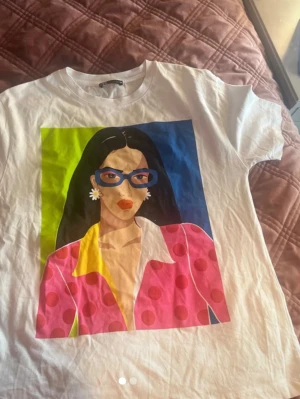 Vit t-shirt med färgglatt tryck - Vit t-shirt med ett stort färgglatt tryck framtill som föreställer en tecknad person i rosa prickig kavaj och gul skjorta. T-shirten har rund hals och korta ärmar. Perfekt för dig som gillar statement-plagg och grafiska motiv.