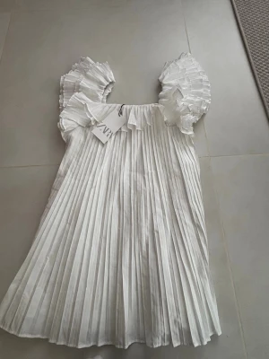 Vit plisserad klänning från Zara MED PRISLAPP - Supersöt helt ny! vit kortklänning från Zara med plisserad design och stora volangärmar. Klänningen har en luftig passform och är perfekt för sommarens alla tillfällen. Tillverkad i ett lätt material som ger ett elegant fall. Prislapp kvar 