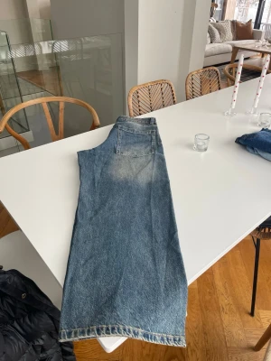 Blå raka jeans med vintagekänsla - Säljer ett par blå jeans med rak passform och klassisk femficksdesign. Jeansen har en snygg tvättad look med ljusare partier och är tillverkade i denim. Perfekta för en avslappnad och trendig stil.