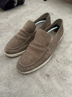 Bruna mockaloafers från Mazzeltov - Snygga loafers i brun mocka från Mazzeltov med klassisk siluett och vit sula. Skorna har en stilren design utan snörning och passar perfekt till en avslappnad men ändå uppklädd look. Mjukt material och diskreta sömmar ger en clean vibe.