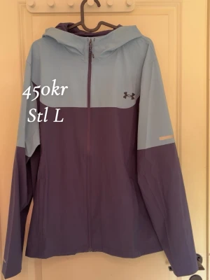 Blå och lila vindjacka Under Armour - Snygg vindjacka från Under Armour i tvåfärgad design med ljusblå upptill och lila nedtill. Jackan har huva, hel dragkedja framtill och logga på bröstet. Perfekt för blåsiga dagar och sportiga outfits.