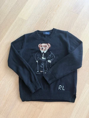 Svart stickad tröja med björnmotiv RL - Stickad svart tröja från Polo Ralph Lauren med ikoniskt björnmotiv på bröstet och RL-broderi vid nederkanten. Tröjan har rund halsringning, långa ärmar och ribbade muddar. Perfekt för dig som gillar klassiska och lekfulla detaljer. Använd 2-3 gånger hör av dig vid frågor pris kan diskuteras
