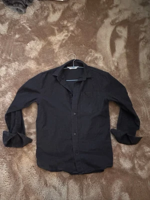 Blå skjorta - Klassisk svart skjorta med lång ärm och knappar framtill. Skjortan har en enkel bröstficka och är tillverkad i mjuk bomull. Perfekt att styla med jeans eller chinos för en clean look.
