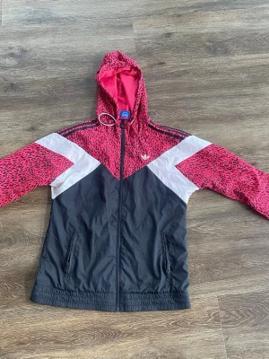 Adidas vindjacka - stl 36 - Snygg vindjacka från adidas med rosa leopardmönster på huva och ärmar, svarta partier och vita detaljer i V-form. Jackan har dragkedja framtill, två fickor och elastisk nederkant. Perfekt för dig som vill sticka ut med sportig stil.