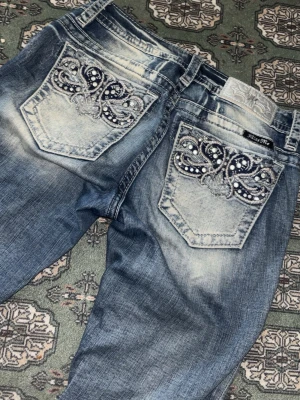 Blå Miss Me jeans med broderade fickor - Säljer ett par blå Miss Me jeans med snyggt slitna detaljer och raka ben. Bakfickorna har broderade mönster med pärlor och paljetter som ger extra bling. Klassisk femficksmodell i denim med cool tvätt och kontrastsömmar.