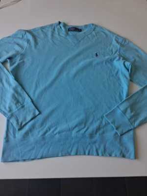 Ljusblå tröja från Polo Ralph Lauren - Snygg ljusblå långärmad tröja från Polo Ralph Lauren med klassisk rund halsringning och broderad logga på bröstet. Tröjan är i mjukt material och har ribbade muddar vid ärmslut och nederkant. Perfekt för en chill och stilren look. Varan är i mycket bra skick i storlek M. Hör av dig vid frågor