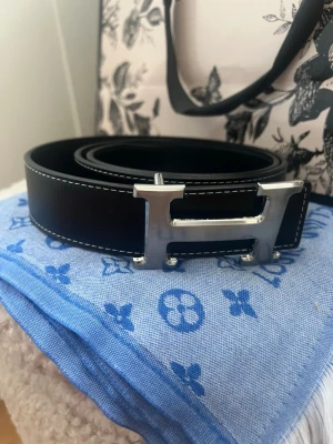 Svart skärp med H-spänne - Snyggt svart skärp med det ikoniska H-spännet i silverfärgad metall. Skärpet har vit kontrastsöm längs kanterna och är tillverkat i äkta skinn. Perfekt accessoar för att lyfta vilken outfit som helst.
