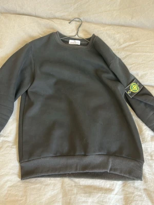 Svart sweatshirt från Stone Island - Cool svart sweatshirt från Stone Island med klassisk logopatch på ena ärmen. Tröjan har rund halsringning och ribbade muddar vid ärmslut och nederkant. Perfekt för dig som gillar streetwear och vill ha något stilrent.